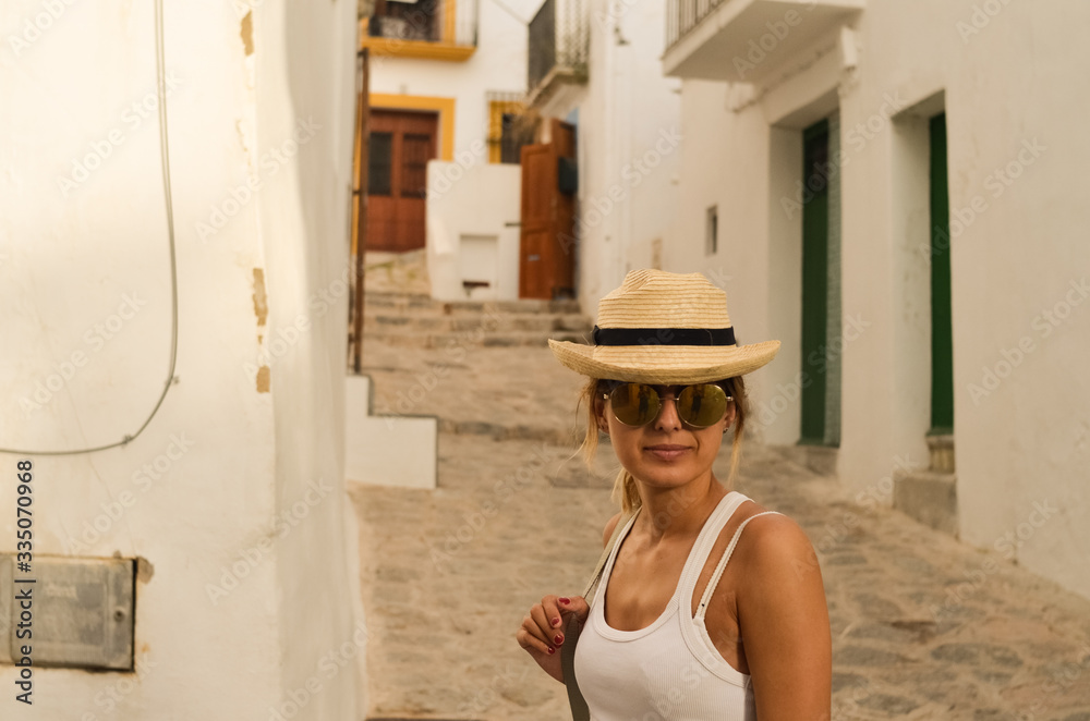 White woman with Straw hat, sunglasses and backpack in a white traditional street in Ibiza