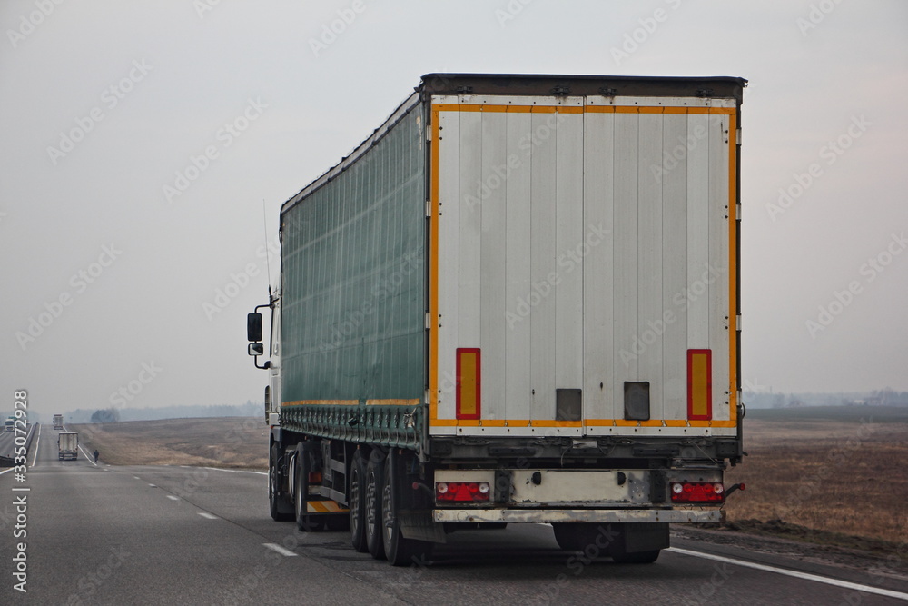 Semi truck van drive on dry suburban asphalt highway at spring day, rear-side view – international logistics, cargo transportation, trucking industry