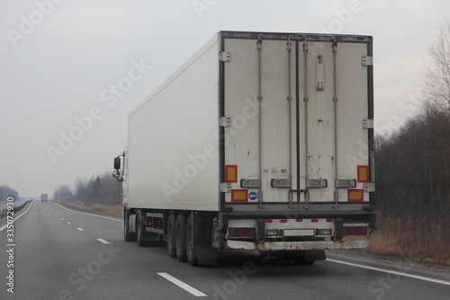 Heavy truck with white semi trailer drive on suburban asphalted road at spring day on gray sky background rear-side view close up – international logistics, cargo transportation, trucking industry