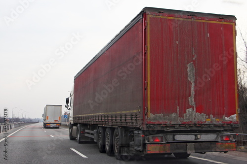 Old red semi truck move on two lane suburban asphalt highway at spring day, rear side view – international logistics, cargo transportation, trucking industry