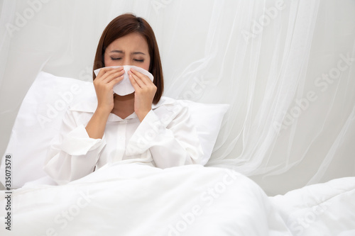 Young Asian woman sneezing in a tissue on the white bed at bedroom, Flu and allergy concept