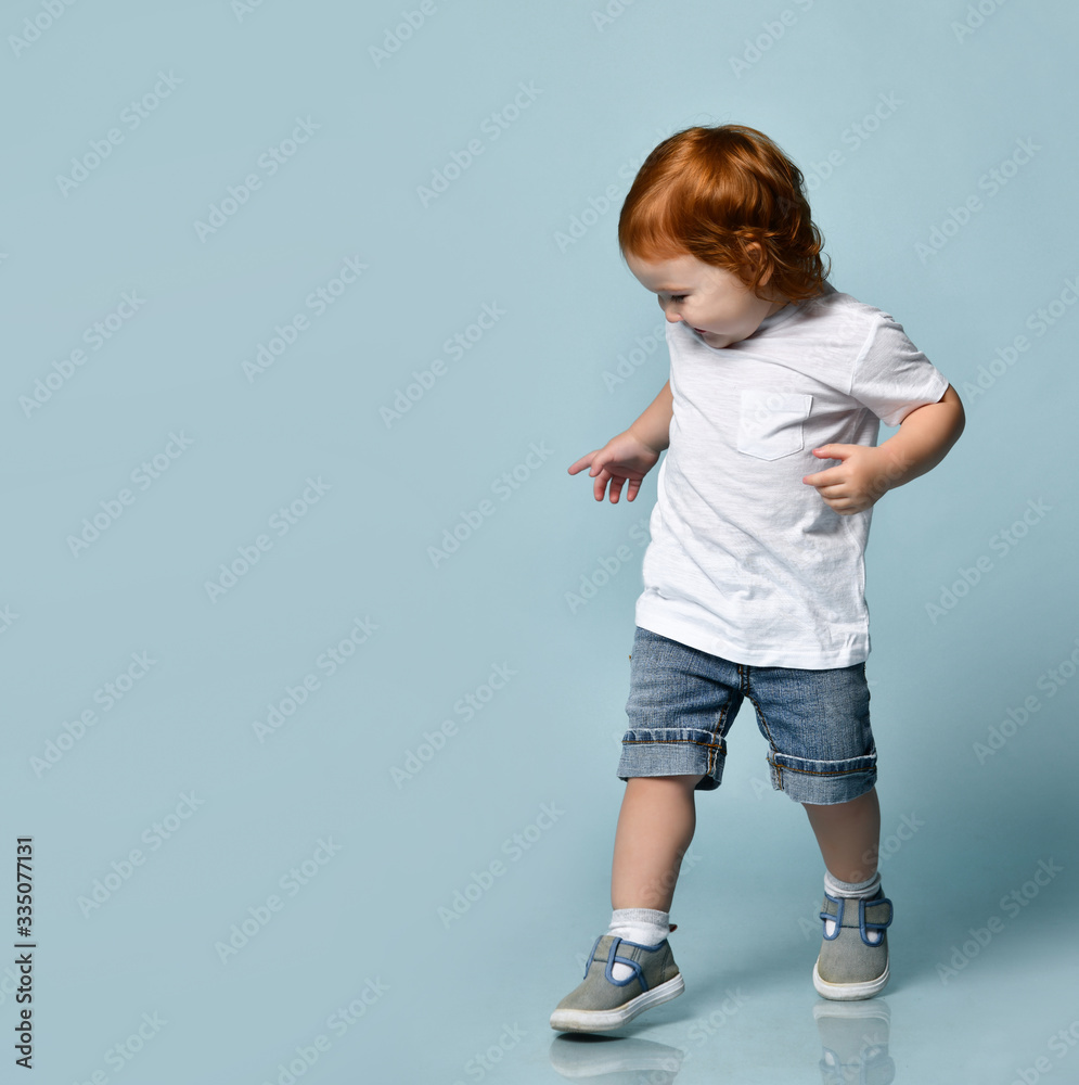 Little ginger toddler boy or girl in white t-shirt, socks and shoes, denim shorts. Child is smiling, walking on blue background