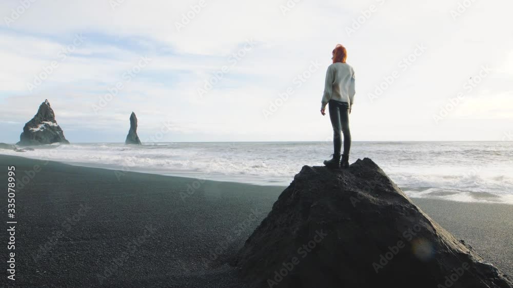 Beautiful red-haired woman traveler standing on a huge stone on volcanic black sand beach in Iceland. Slow motion shot. Reynisfjara Beach around Vik. Troll toes Reynisdrangar mountains on background Beautiful red-haired woman traveler standing on a huge stone on volcanic black sand beach in Iceland. Slow motion shot. Reynisfjara Beach around Vik. Troll toes Reynisdrangar mountains on background