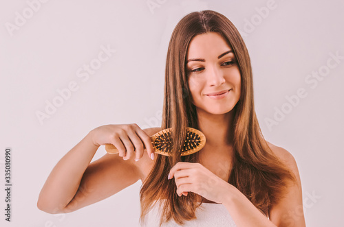 Portrait of brunette young beautiful woman brushing glossy straight hair and smiling isolated. Attractive happy girl combing shiny healthy hair with wooden brush. Hair care routine, natural beauty