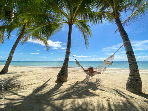 Girl resting and swinging on hammock in paradise resort.Beautiful sandy white beach with turquoise sea in grove with palm trees. Shadows on sand