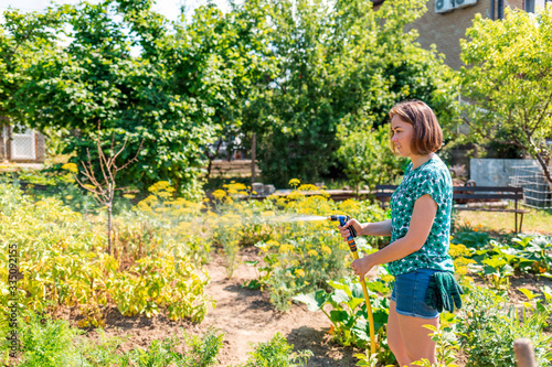 A woman in casual clothes and gloves in her pocket, watering a garden with greenery with a hose. Home garden on the background.The concept of gardening and summer recreation