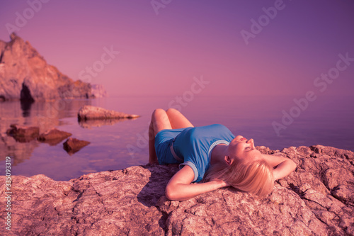 Young relaxed caucasian woman lay on a stone and enjoys the rays of the summer sun on the background of the sea during the long-awaited vacation on a summer day. Rest and recovery concept