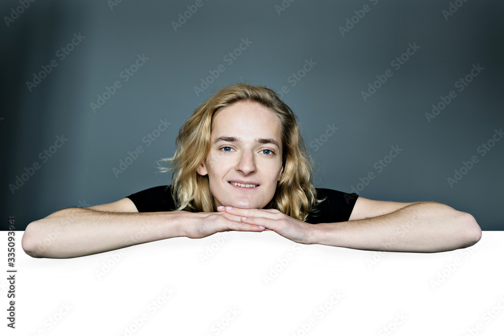 A young attractive man peeks out from above a white wall on a gray background. He laid his head in his hands. The elbows are apart. A young attractive man peeks out from above a white wall on a gray background. He laid his head in his hands. The elbows are apart.