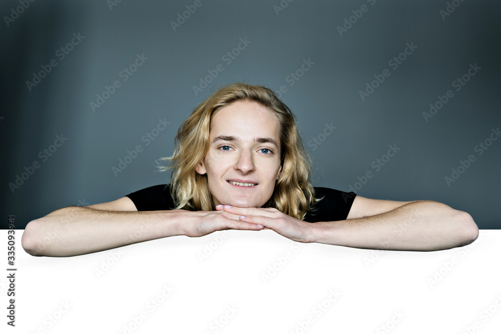 A young attractive man peeks out from above a white wall on a gray background. He laid his head in his hands. The elbows are apart.