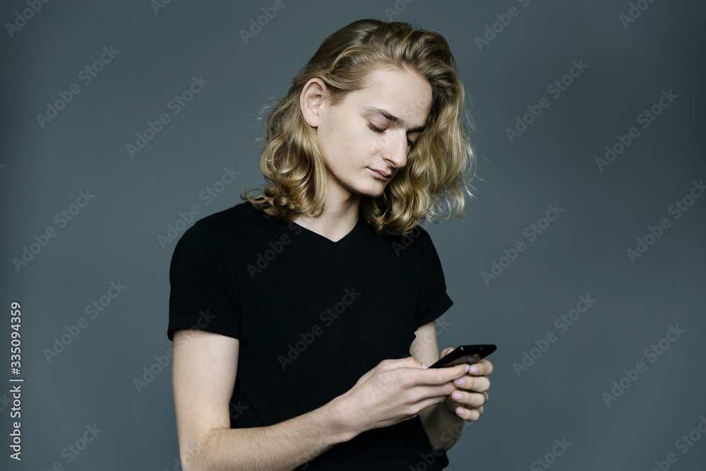 Young man with long blonde hair taking pictures of something with a phone with a strained expression on his face on a black background Young man with long blonde hair taking pictures of something with a phone with a strained expression on his face on a black background