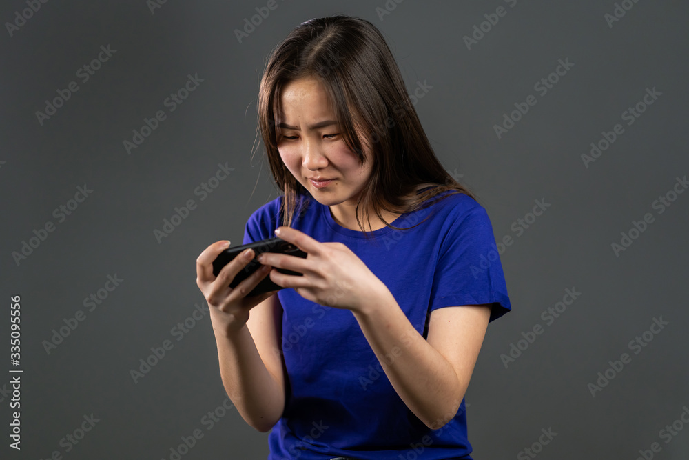 Girl with asian appearance playing game on smartphone on grey studio wall. Using modern technology - apps, social networks