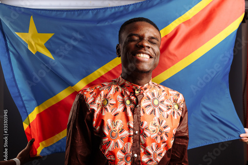 A black african man smiling and posing before the flag of the Democratic Republic of the Congo.