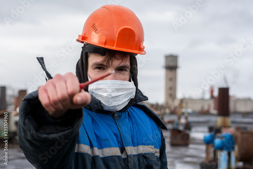 a working fitter in a protective helmet holds a red turn screw in his outstretched hand. The main focus on the turn screw. On the face a protective mask. Action takes place at the factory