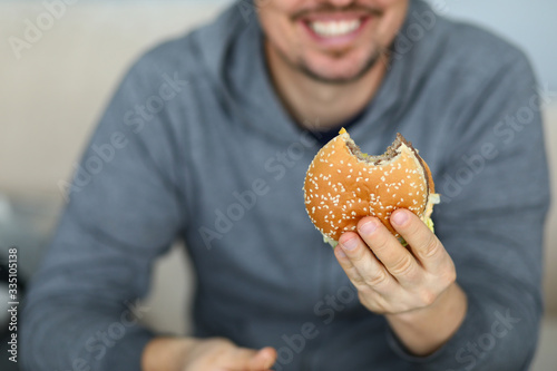 Focus on male hand holding big tasty hamburger full of mischievous calories and harmful cholesterol. Happy and fat person consuming portion of junk food