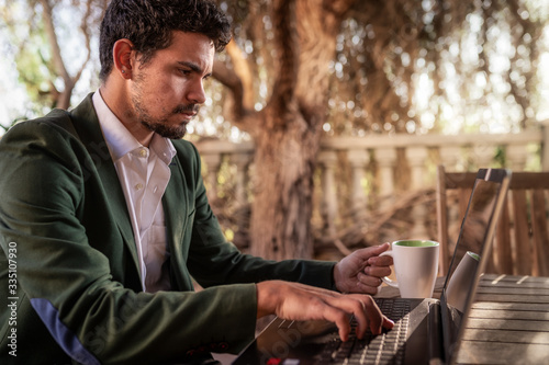 primer plano.Hombre joven con chaqueta verde sentado en el jardin, trabaja concentrado en su ordenador portatil mientras bebe un cafe. Homework. 