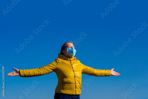 girl in a medical mask with arms spread apart on the sides enjoying fresh air in freedom against a blue sky with a copy space. © Александр Беспалый