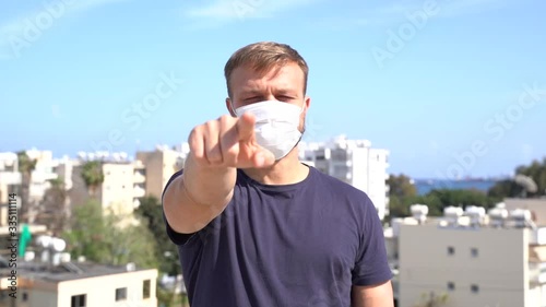 Portrait of caucasian white male, millennial, in medical face mask showing on directly to the camera, pointing hand signal. Coronavirus world pandemic concept of social distance and responsibility