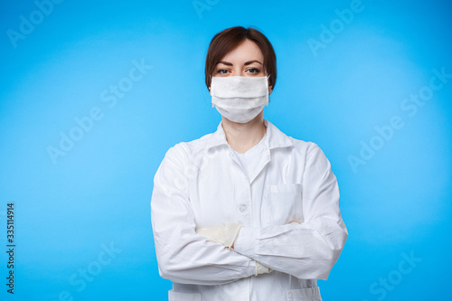 Portrait of professional female doctor in white uniform and protective mask posing isolated. Practitioner woman medical personnel in safety suit stand with crossed hands on blue studio background