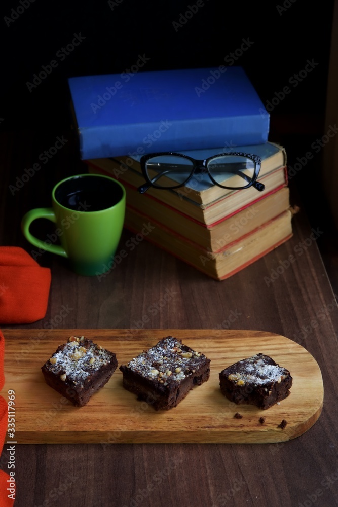 Three chocolate brownie sprinkled with castor sugar on a wooden board in the foreground with a green mug and a pile of books in the background.  A pair of glasses on the stack of books.