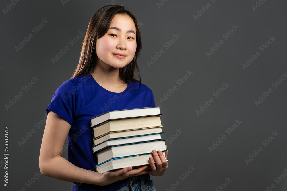 Asian student on grey background in studio holds stack of university books from library. Woman smiles, she is happy to graduate. Asian student on grey background in studio holds stack of university books from library. Woman smiles, she is happy to graduate.