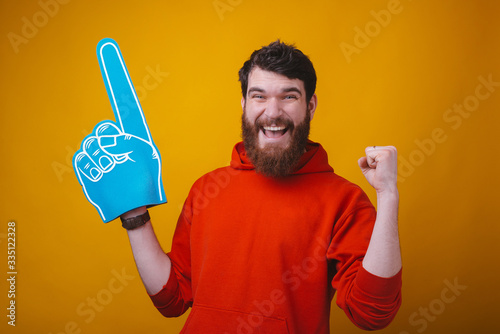 I am number one fan. Photo of a bearded man is wearing a blue foam fan glove on yellow background. © Vulp