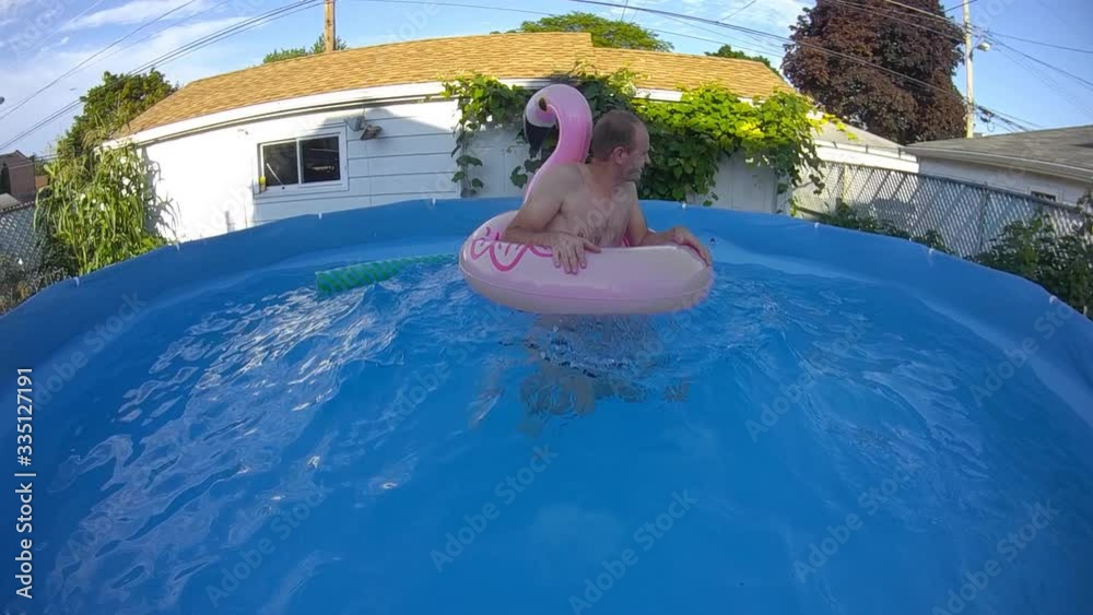 Slow motion of a man in inflatable floating flamingo tube having fun in back yard swimming pool while floating up and down on volcano wave.
