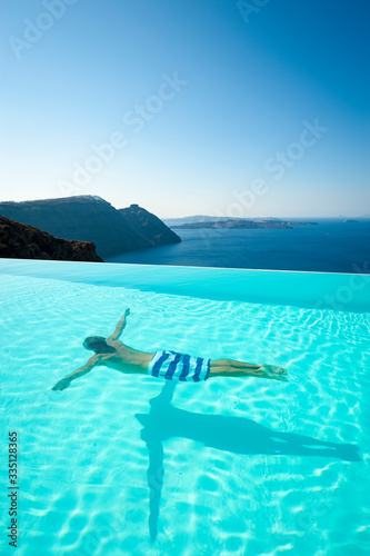 Dramatic caldera view of unrecognizable man swimming underwater in turquoise infinity pool overlooking the Mediterranean Sea in Santorini, Greece
