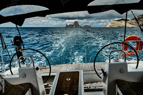 The view of the sea and mountains from the sailboat, edge of a board of the boat, oat stern, steering wheel, without people, slings and ropes, splashes from under the boat, sunny weather, dramatic sky