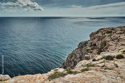 The Landscape of the balearic sea and improbable mountains, azure water, the storm sky, lonely buildings at tops of mountains, sail boat is on background