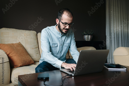 Young man working online with a laptop sitting on the sofa in the afternoon at home