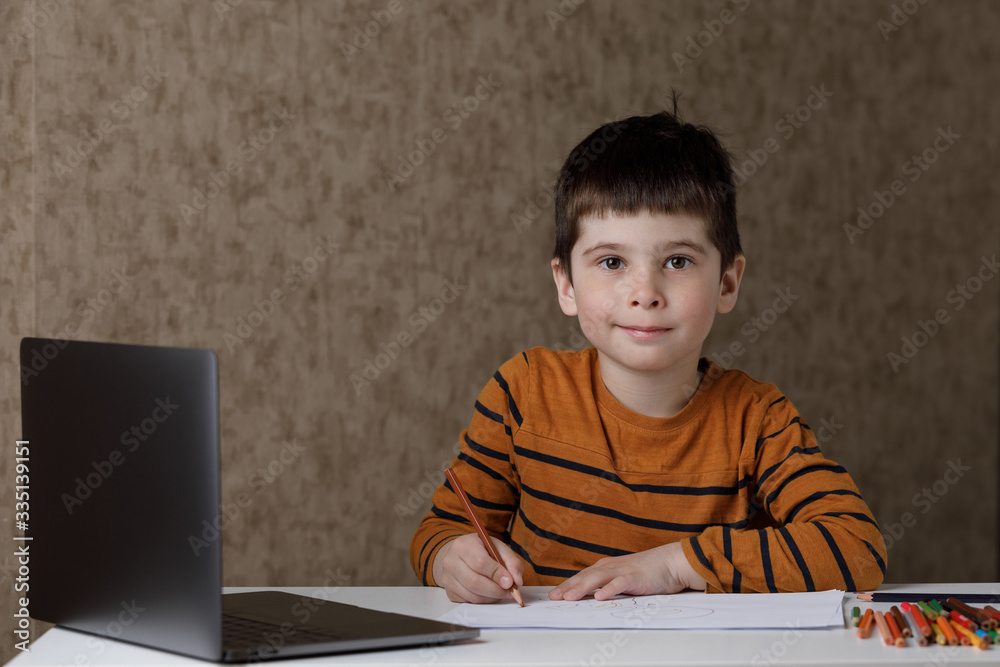 the child learns remotely through the computer. The boy draws with pencils and looks into the laptop the child learns remotely through the computer. The boy draws with pencils and looks into the laptop