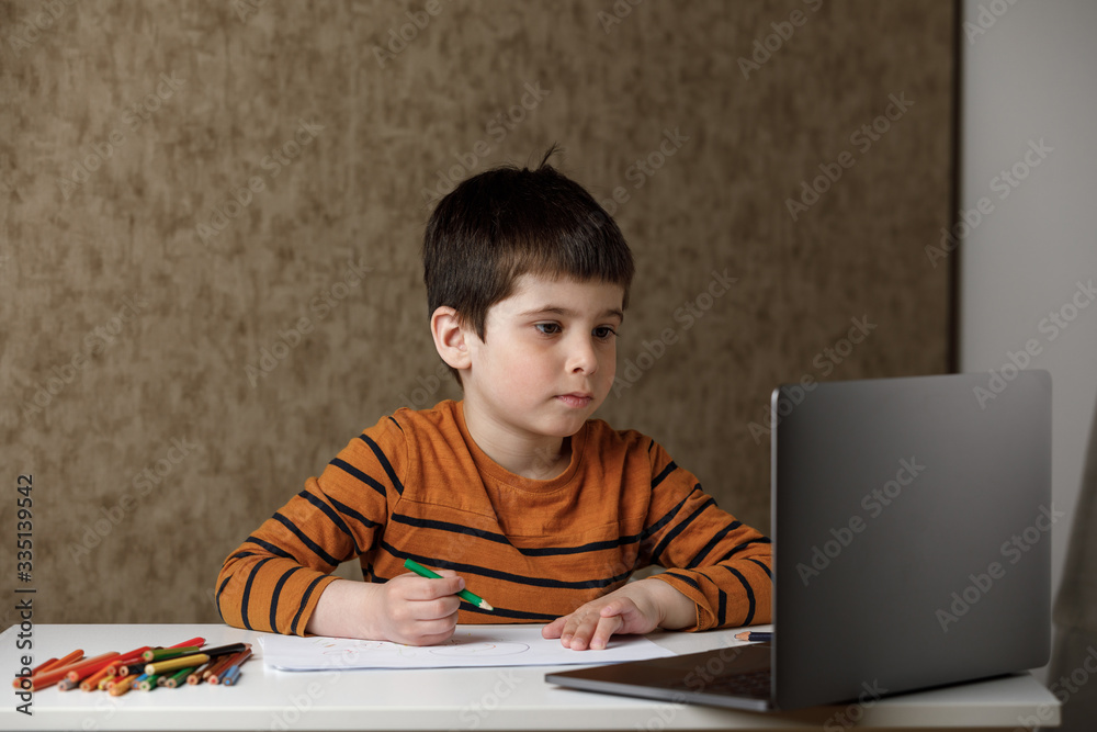 The boy draws with pencils on paper and looks into the laptop. Remote training via the Internet The boy draws with pencils on paper and looks into the laptop. Remote training via the Internet