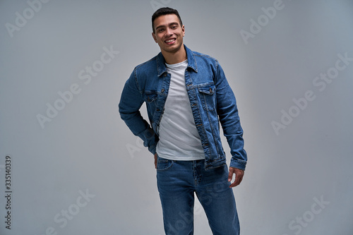 Happy guy in a t-shirt in a denim jacket stands with his hand in the back pocket of the jeans. Fashion concept