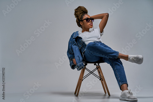 Confident lady in sunglasses in white t-shirt in jeans with denim jacket posing on camera sitting on chair with leg lies on the knee. Fashion concept