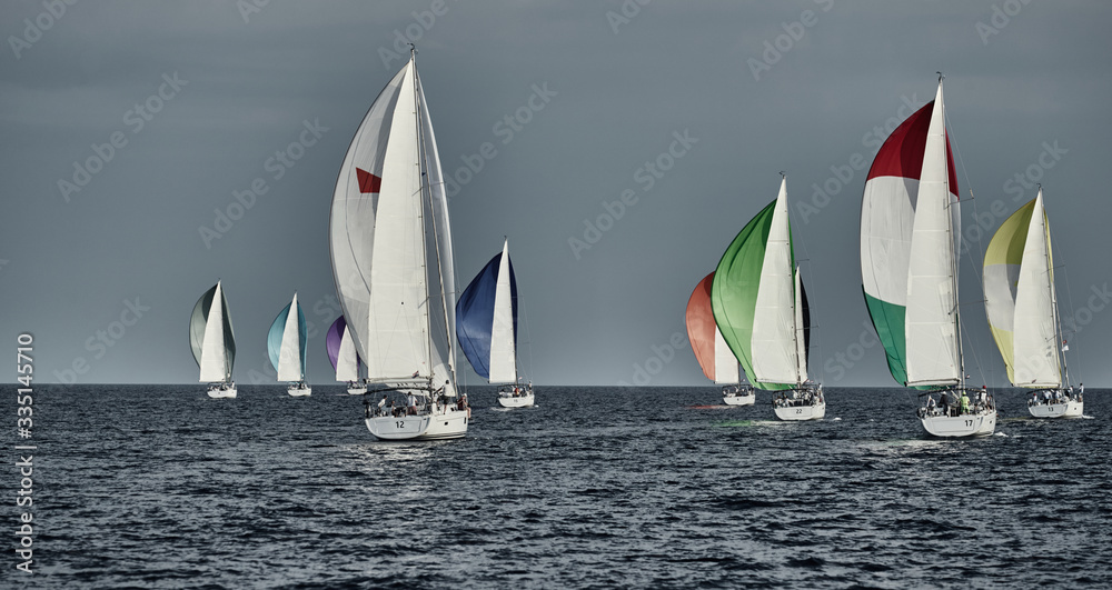 Fototapeta premium Sailboats compete in a sailing regatta at sunset, sailing race, reflection of sails on water, multi-colored spinaker, boat number aft boats, big white clouds,