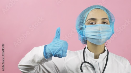 Female doctor in medical face mask and medical gloves showing handgesture thumbs up. Dont worry, be happy. After covid-19 or post-pandemic period. Celebrating victory over coronavirus photo