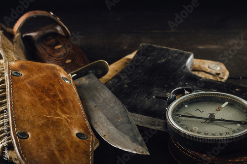 Old military compass, rusty hunting bushcraft knife, and small axe on the dark wooden table. Leather cases, close up, front view, survival hunting concept. photo
