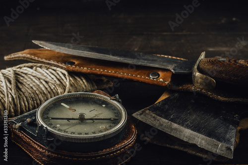 Old military compass, rusty hunting bushcraft knife, small axe and a linen rope on the dark wooden table. Leather cases, front view, survival hunting concept.