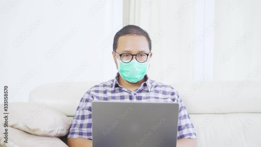 Closeup of man working with laptop while wearing a mask and sitting on the sofa in the living room at home Closeup of man working with laptop while wearing a mask and sitting on the sofa in the living room at home