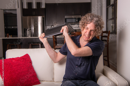 A man works from home on a laptop computer, in a city apartment, with a modern kitchen behind. He's frustrated and wants to throw his laptop. photo