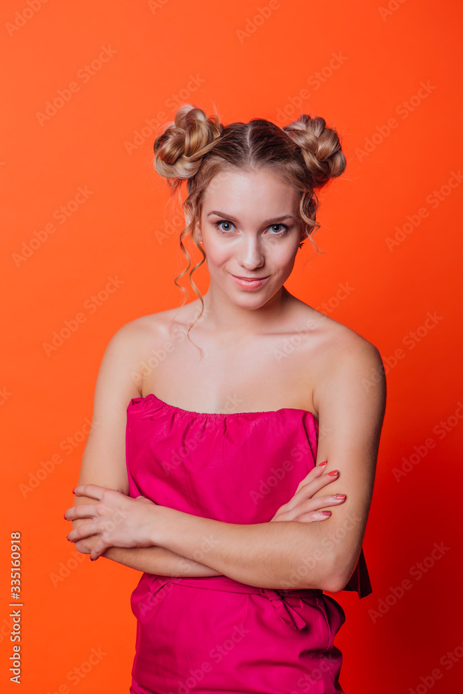 The girl in a red dress on a orange background in the studio. Blonde girl with two hair knots looking to the camera and smiling.