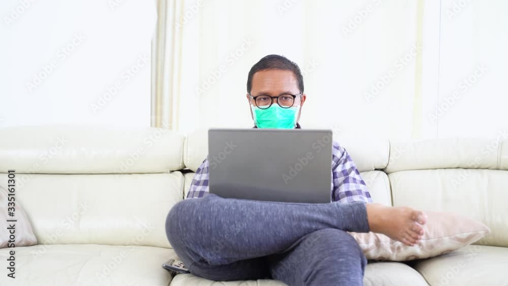 Young man wearing a mask to prevent corona virus while working with laptop on the sofa at home. Shot in 4k resolution
