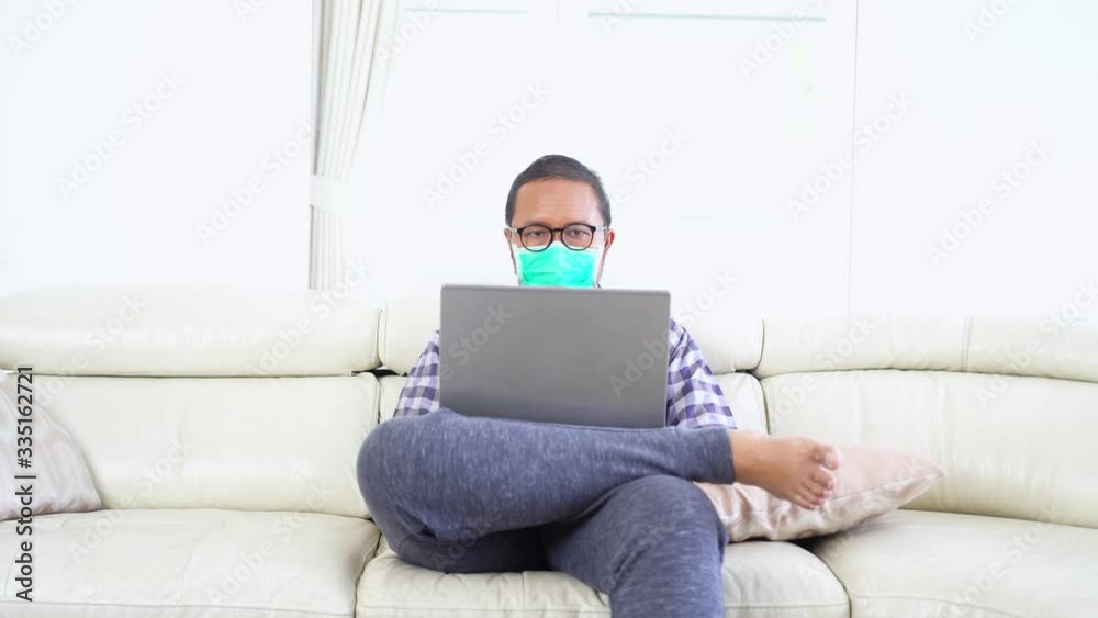Young man working at home to prevent corona virus with a laptop computer while sitting on the sofa and wearing a mask. Shot in 4k resolution Young man working at home to prevent corona virus with a laptop computer while sitting on the sofa and wearing a mask. Shot in 4k resolution
