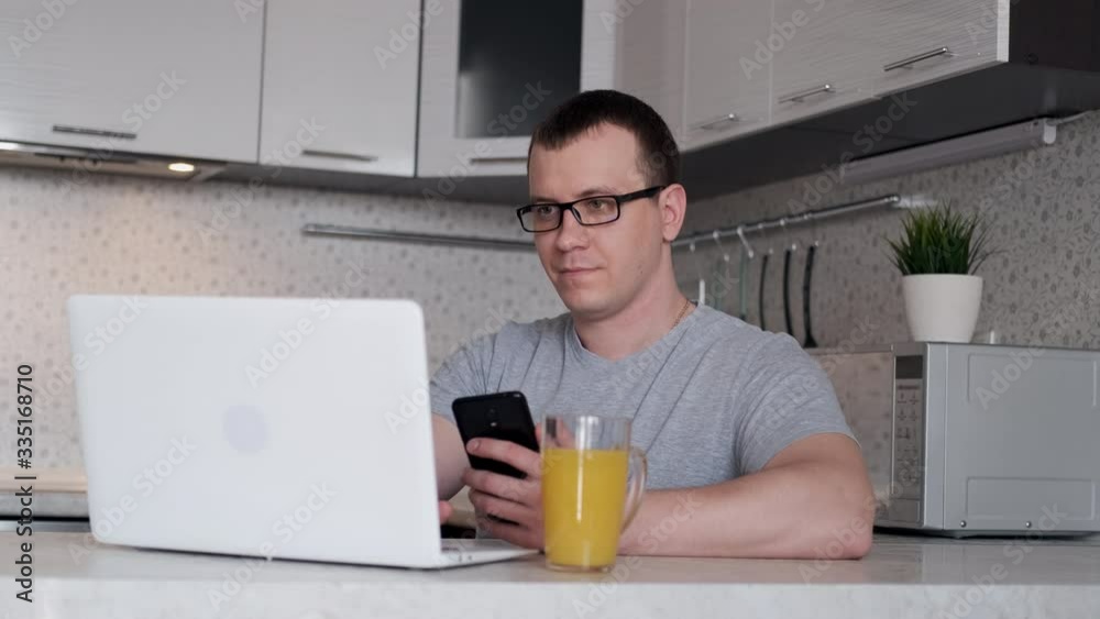 A male freelancer works from home using a phone and a laptop while sitting at a table. A male freelancer works from home using a phone and a laptop while sitting at a table.