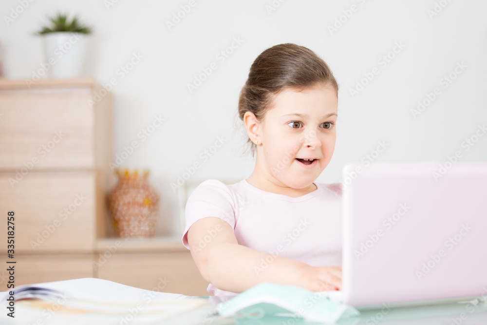 Delighted little girl with a pink computer, a laptop is sitting on an online training, a happy schoolgirl is delighted with distance learning online. Online concept of school education . Delighted little girl with a pink computer, a laptop is sitting on an online training, a happy schoolgirl is delighted with distance learning online. Online concept of school education .