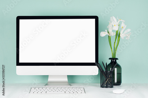 Elegant spring home workplace with blank computer screen, black stationery, keyboard, mouse, white fresh flowers in vase in green mint menthe interior on white wood desk.
