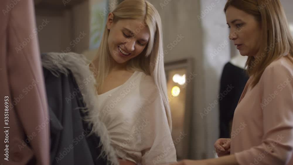 Handheld shot of happy blond woman and her mother looking at dress with fringe in boutique and discussing it Handheld shot of happy blond woman and her mother looking at dress with fringe in boutique and discussing it