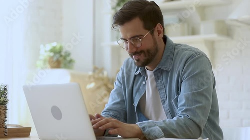 Concentrated young man freelancer in eyeglasses looking at laptop screen, working remotely at home. Head shot focused smiling guy chatting in social network, typing message, internet web surfing.