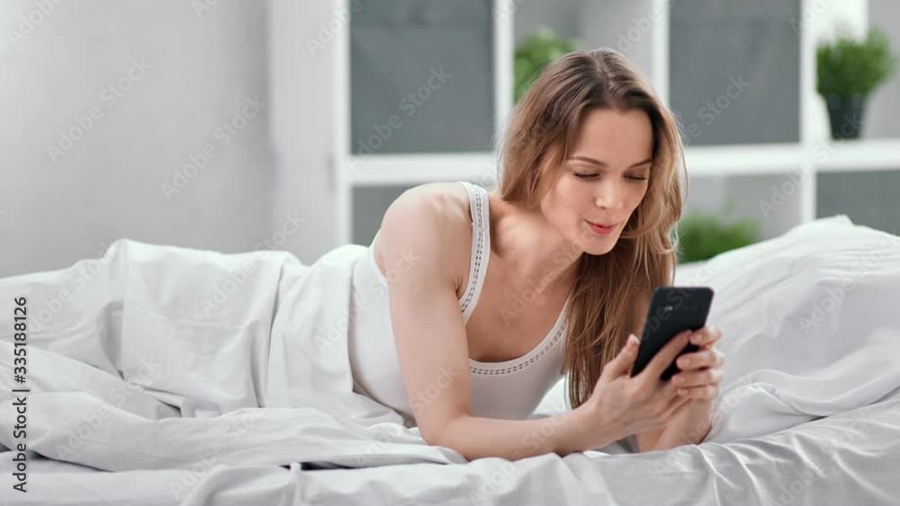 Smiling young woman lying on bed relaxing chatting using smartphone. Shot with RED camera in 4K Smiling young woman lying on bed relaxing chatting using smartphone. Shot with RED camera in 4K