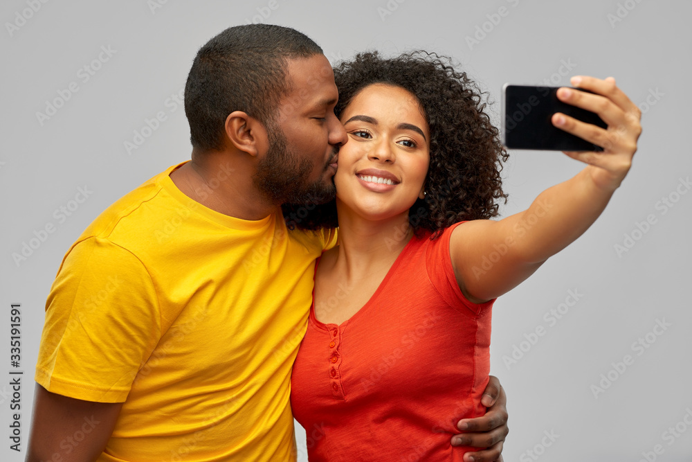 Obraz premium technology and people concept - happy smiling african american couple taking selfie by smartphone and kiss over grey background